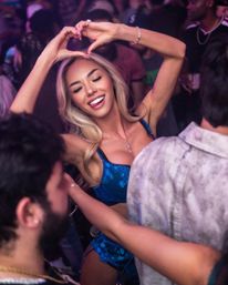 Smiling woman in blue lingerie making a heart with her hands on a crowded nightclub dance floor under colorful neon lights