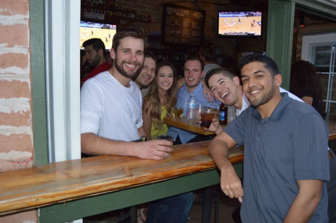 Cheerful group of six friends leaning at a wooden bar window in a sports bar, smiling and holding drinks with TVs showing a basketball game in the background.