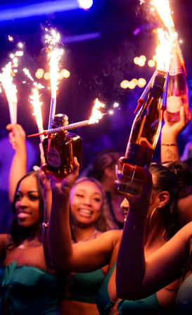 VIP nightclub party with bottle service and sparklers on champagne and liquor bottles, energetic crowd celebrating late-night