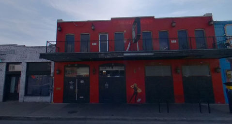 Bright red two-story storefront on a city street with an upper balcony, black awning, closed metal roll-up doors and a small painted mural of a stylized woman, next to a white-brick shop.