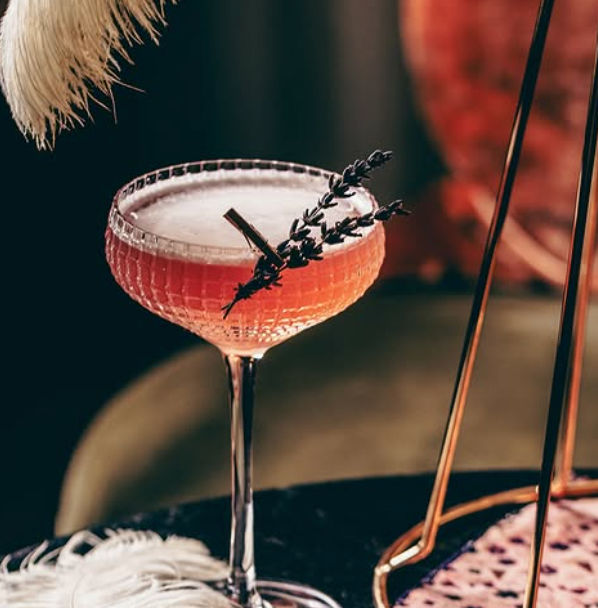 Pretty pink craft cocktail in a vintage coupe glass topped with foam and a lavender sprig, set on a dark table with gold-accented bar decor.