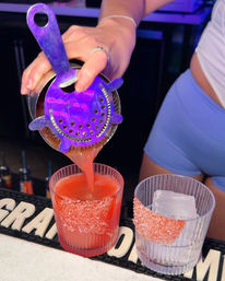 Close-up of a shaker pouring a bright red cocktail through a purple-lit strainer into a pink-salt-rimmed rocks glass, next to a second glass with a jumbo ice cube on a neon-lit bar counter.