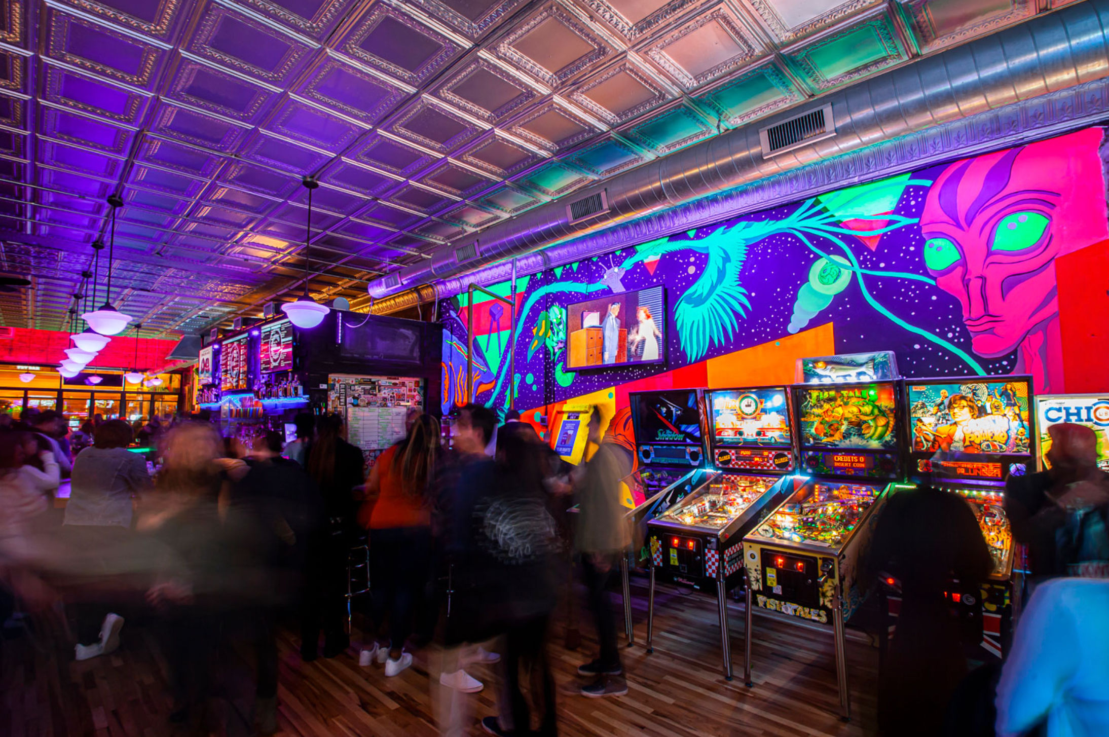 Neon-lit arcade bar interior with a row of illuminated pinball machines, vibrant psychedelic mural and embossed tin ceiling, hanging pendant lights and a lively blurred crowd enjoying nightlife.