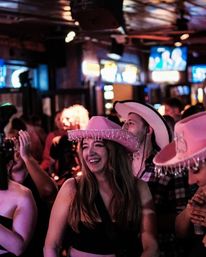Young woman laughing in a crowded bar wearing a pink fringed cowboy hat, surrounded by people in cowboy hats under neon lights — lively country-themed nightlife scene