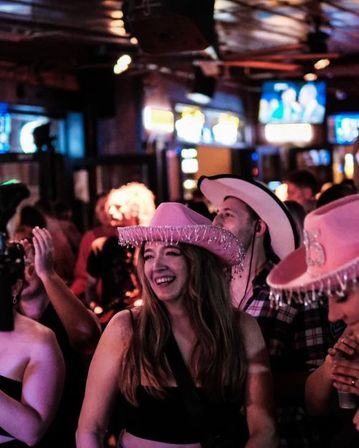 Young woman laughing in a crowded bar wearing a pink fringed cowboy hat, surrounded by people in cowboy hats under neon lights — lively country-themed nightlife scene