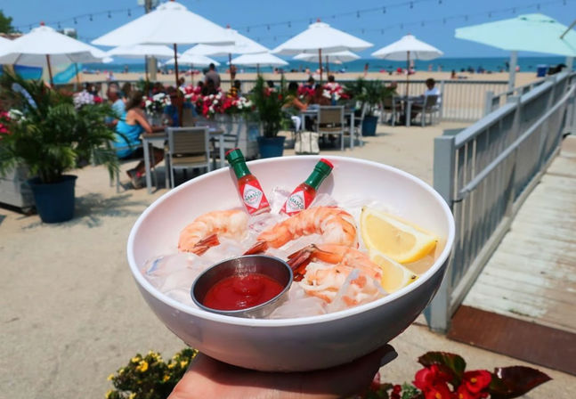 Beachside shrimp cocktail on ice with lemon wedges and mini hot sauce bottles held over a sunny oceanfront patio and boardwalk with white umbrellas and outdoor dining