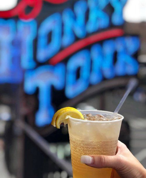Hand holding a plastic cup of refreshing iced lemon drink with a straw and lemon wedge, bubbly amber liquid in front of a blurred neon sign on an outdoor urban patio.
