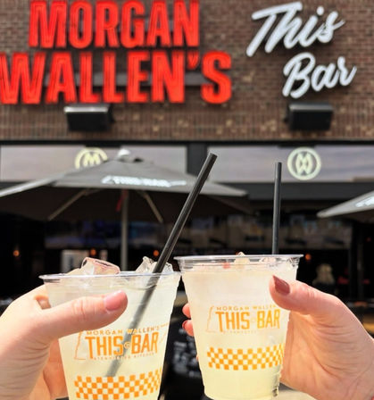 Two hands clinking plastic cups of icy lemonade with black straws on a sunny outdoor bar patio, brick storefront and umbrellas with a glowing neon sign in the background.