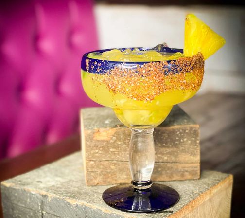 Icy pineapple margarita in a blue-rimmed glass with chili-salt rim and pineapple wedge, set on stacked wooden blocks against a pink tufted backdrop
