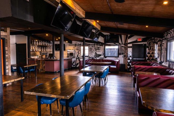 Inviting industrial-chic restaurant interior with warm wooden floors, long communal wood tables and blue chairs, red leather booths, graphic black-and-white wall mural, overhead flat-screen TVs and a bar with hanging glassware.