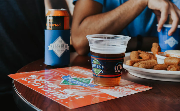 Plastic cup of beer on a wooden bar table next to a plate of fried appetizers and a canned drink in a blue koozie, casual pub snack scene.