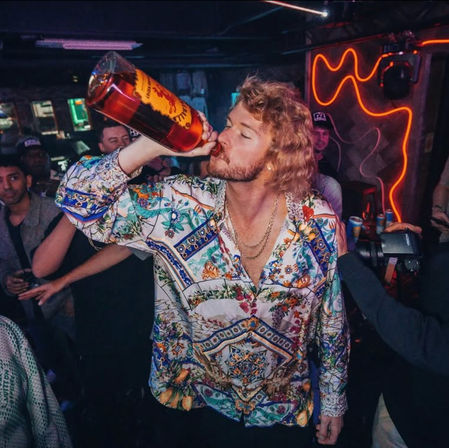 Man in a colorful patterned shirt chugging from a large liquor bottle in a neon-lit nightclub crowd, lively bar party atmosphere.