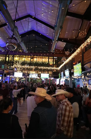 Crowded indoor bar with twinkling string lights and disco ball under a high industrial ceiling; patrons in cowboy hats watch sports on multiple TVs.