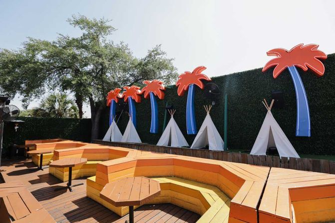 Tropical-themed outdoor patio with orange and yellow tiered wooden benches and hexagonal tables on a deck, white teepee tents and oversized neon palm signs along a hedge wall.