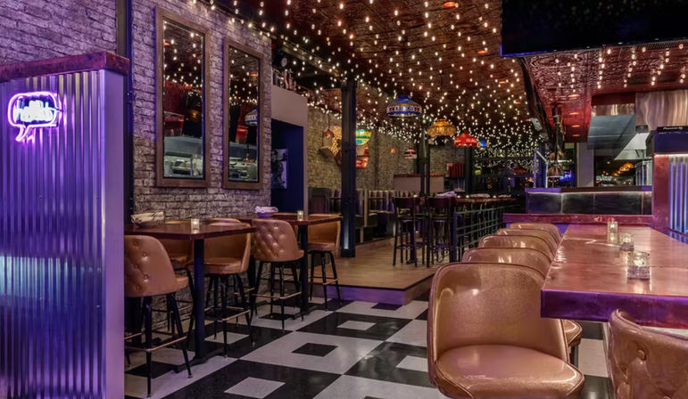 Industrial-chic bar interior with exposed brick walls, checkerboard tile floor, leather bar stools, wooden tables, colorful pendant lamps and overhead string lights creating a cozy evening dining atmosphere.