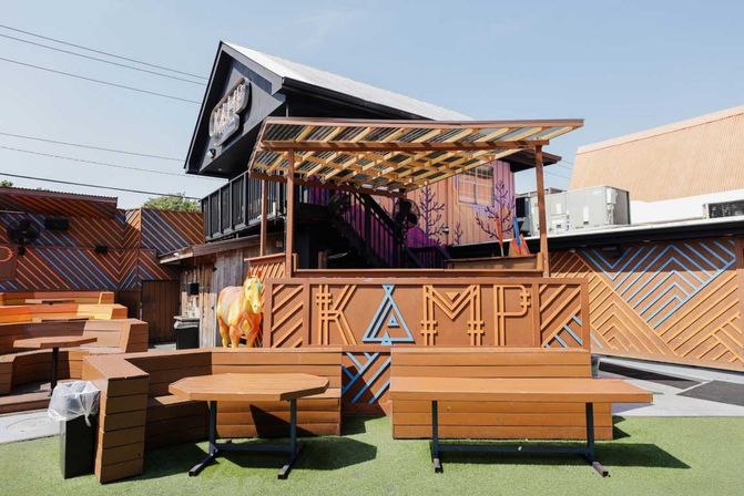 Sunny outdoor patio bar with tiered wooden benches and round tables on green turf, pergola-covered stage with chevron wood paneling and a quirky painted cow statue
