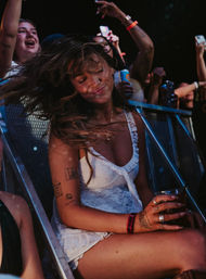 Woman dancing at a nighttime music festival near the front-row barrier, hair flying and eyes closed, smiling while holding a drink as fans cheer and record on phones.