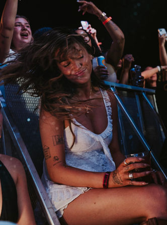 Woman dancing at a nighttime music festival near the front-row barrier, hair flying and eyes closed, smiling while holding a drink as fans cheer and record on phones.