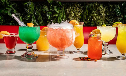Row of colorful tropical cocktails and mocktails on a bar counter — large pink punch bowl exhaling dry-ice fog, bright green margarita with syringe garnish, mason-jar drink, orange and lime slices, salted rims, and lush plant wall backdrop.
