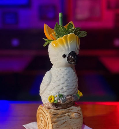 Ceramic white cockatoo cocktail mug perched on a log base, filled with a tropical drink garnished with orange wedges and fresh mint against a neon-lit bar backdrop.