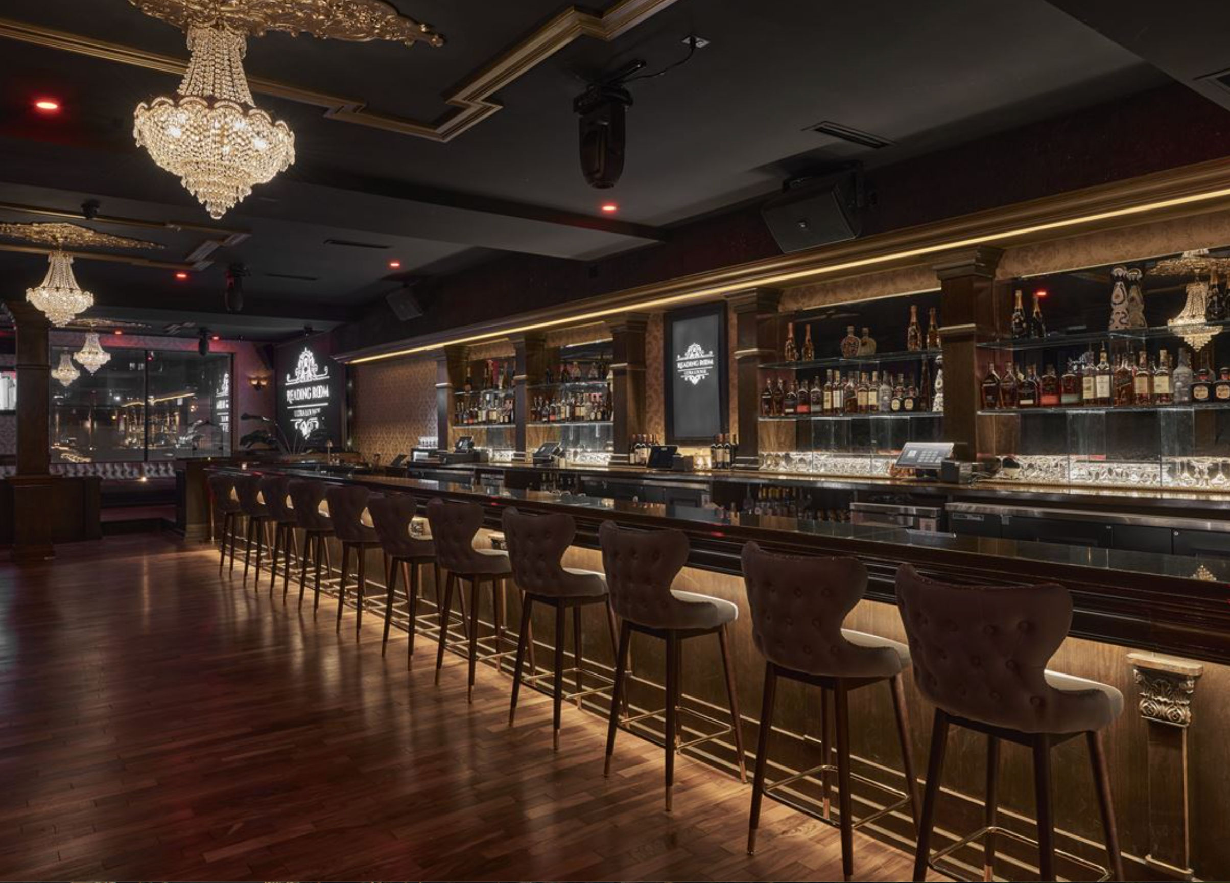 Upscale cocktail lounge interior with crystal chandeliers, long backlit wooden bar lined with tufted leather bar stools, mirrored liquor shelves and warm ambient lighting over polished hardwood floors.