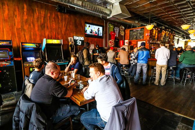 Busy neighborhood arcade bar interior with wood-paneled walls, vintage arcade machines and patrons socializing at tables and the bar.