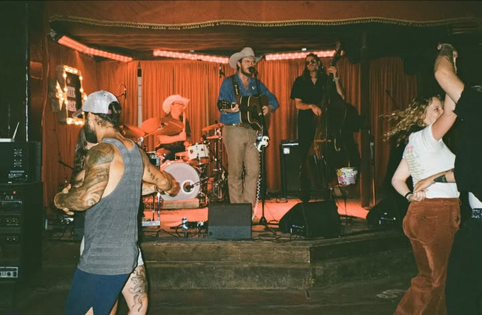 Couples dancing on a wooden floor in front of a small bar stage where a country band plays — singer in a cowboy hat with acoustic guitar, drummer and upright bass under warm red curtains.
