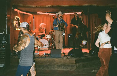 Couples dancing on a wooden floor in front of a small bar stage where a country band plays — singer in a cowboy hat with acoustic guitar, drummer and upright bass under warm red curtains.