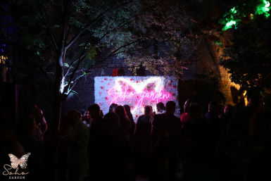 Crowded nighttime garden party — silhouetted guests in front of a flower-covered wall lit by pink neon script and colorful tree lights.