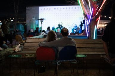 Cozy couple sitting on colorful chairs watching an outdoor jazz concert at night, neon lights on a tree and a live band playing on a lit stage in the background