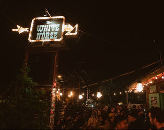 Nighttime urban nightlife scene: a vintage illuminated sign and hanging string lights over a lively outdoor patio crowd on a busy street.