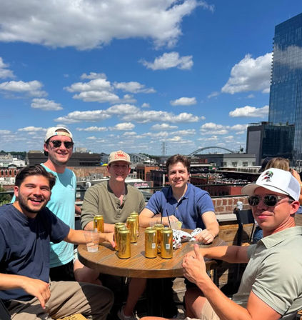 Group of five friends at a sunny rooftop patio bar, golden beer cans on a wooden table, blue sky with fluffy clouds and a riverfront downtown skyline with an arched bridge in the background.