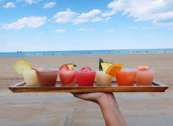 Hand holding a wooden tray of six colorful, fruit‑garnished tropical cocktails on a sandy oceanfront beach under a bright blue sky
