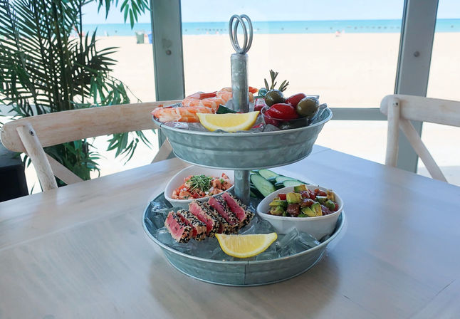 Two-tier galvanized seafood tower on an oceanfront table: ice-filled trays with shrimp cocktail, sesame-crusted seared tuna, avocado poke, marinated olives, cucumber slices and lemon wedges, sandy beach and blue sea beyond.