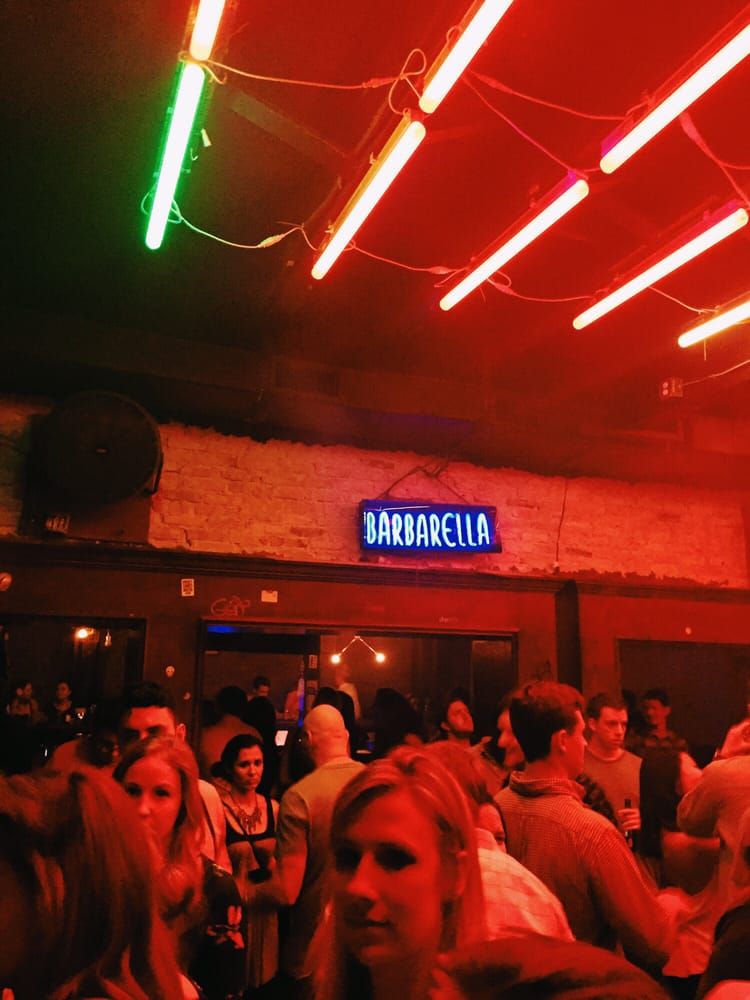 Lively crowded nightclub interior with red neon ceiling tubes, a blue neon sign on exposed brick, and people socializing on the dance floor.