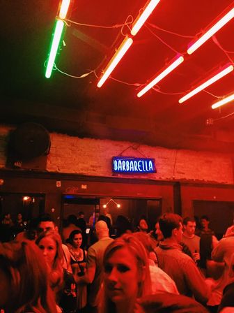 Lively crowded nightclub interior with red neon ceiling tubes, a blue neon sign on exposed brick, and people socializing on the dance floor.
