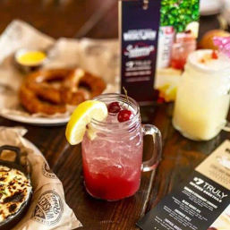 Bright red mason-jar cocktail garnished with cherries and lemon wedge on a rustic restaurant table, with a pretzel and cheesy skillet appetizer in the background