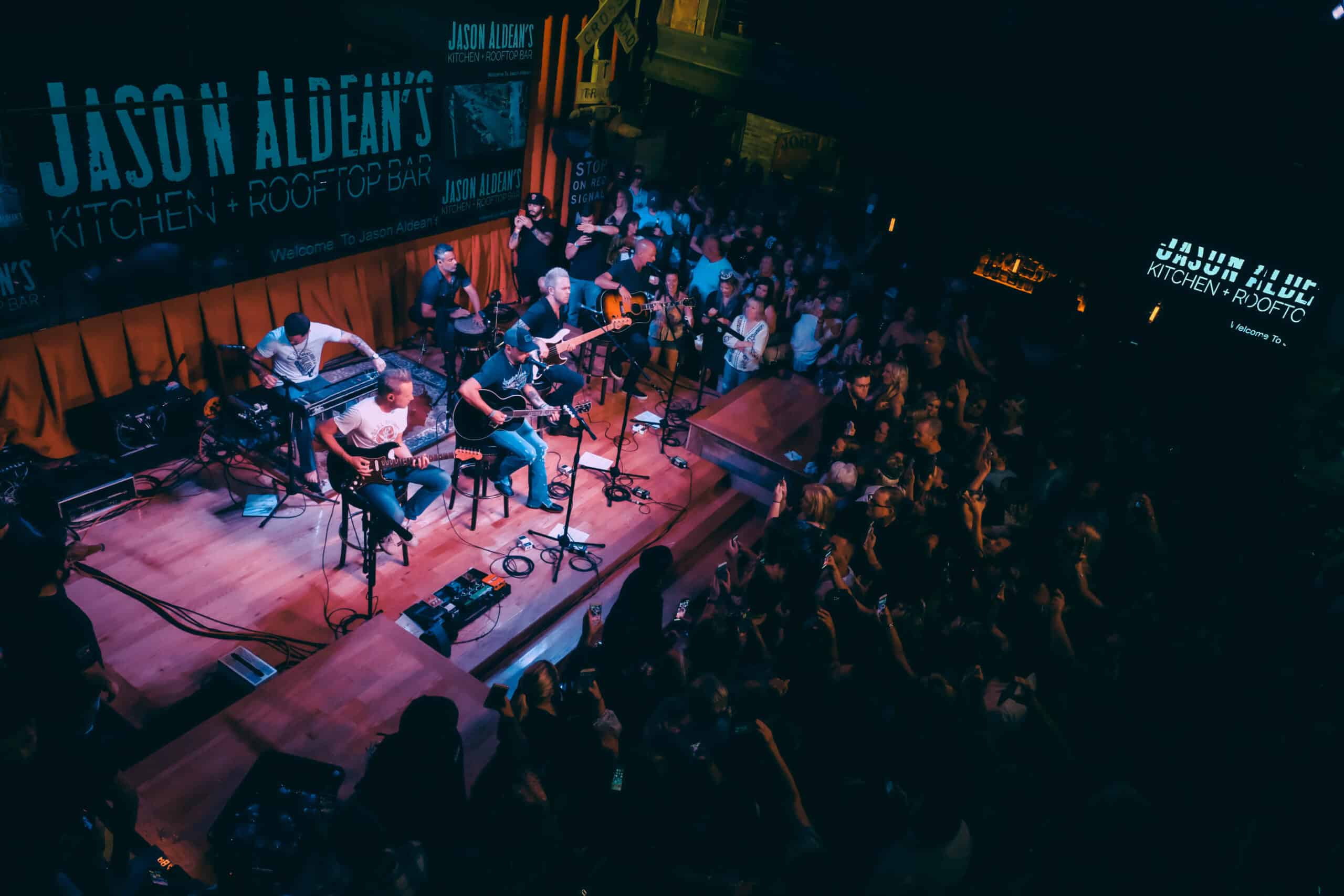 Live acoustic band on a small rooftop-bar stage with guitars and keyboards, performing to a packed, energetic crowd under moody blue lighting.