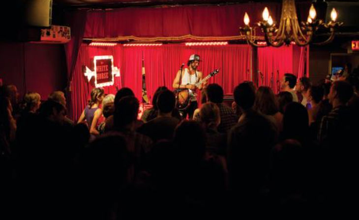 Solo guitarist performing on a small red-curtained stage in an intimate indoor venue, crowd watching under warm chandelier lighting — lively live music nightlife scene.