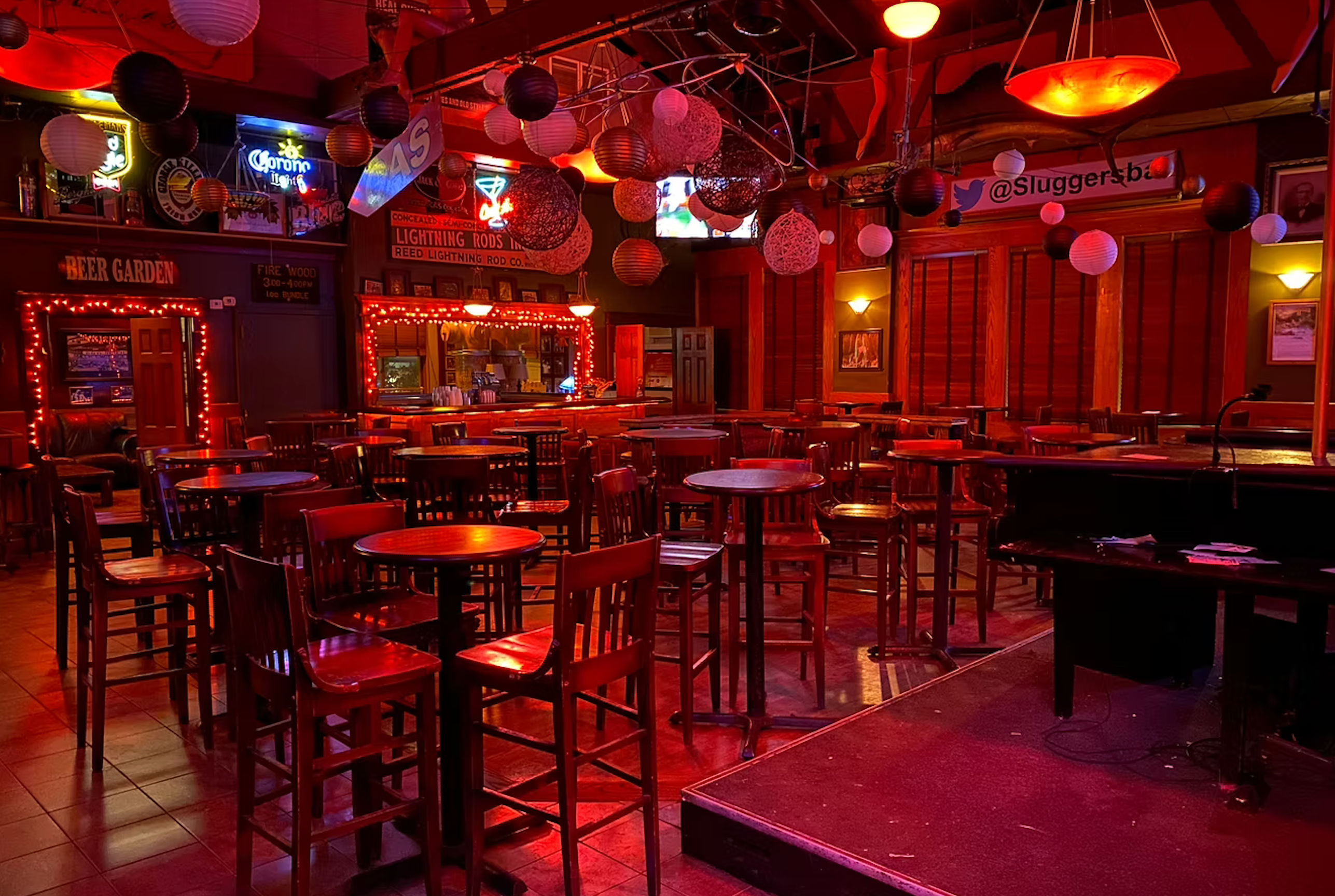 Empty cozy bar interior with warm red lighting, wooden high-top tables and stools, hanging paper lanterns, neon beer signs over a mirrored bar counter, and a small stage for live music