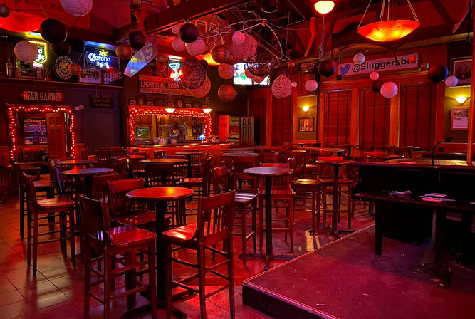 Empty cozy bar interior with warm red lighting, wooden high-top tables and stools, hanging paper lanterns, neon beer signs over a mirrored bar counter, and a small stage for live music