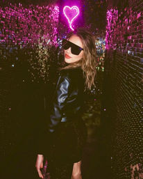 Person in a black leather jacket and oversized sunglasses posing under a pink neon heart against a shimmering sequin wall in a dim, glam nightclub