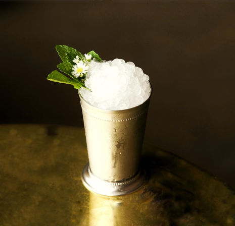 Frosty mint julep-style cocktail in a silver julep cup piled with crushed ice, garnished with mint leaves and tiny white flowers on a brass table against a dark background