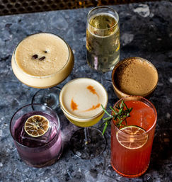 Top-down view of six colorful craft cocktails on a dark marble bar counter — espresso martini with coffee beans, champagne flute, frothy citrus sour, creamy brown cocktail, purple gin drink with dried lime, and pink rosemary spritz.