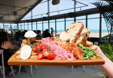 Wooden board with smoked salmon, grilled bread slices, cherry tomatoes, pickled red onions, capers, cream cheese and sliced boiled egg, served on a tray at a sunlit beachfront patio with ocean view