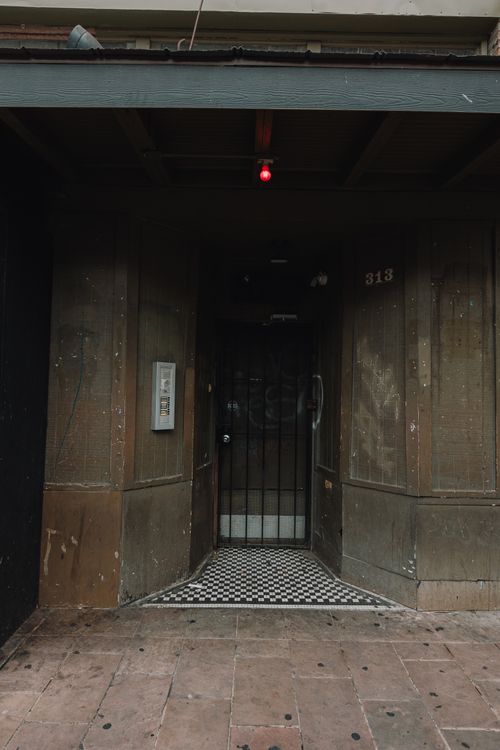Urban storefront entrance with a black metal security gate, worn brown wood facade, black-and-white checkerboard tile threshold, small red hanging bulb above, and scuffed sidewalk.