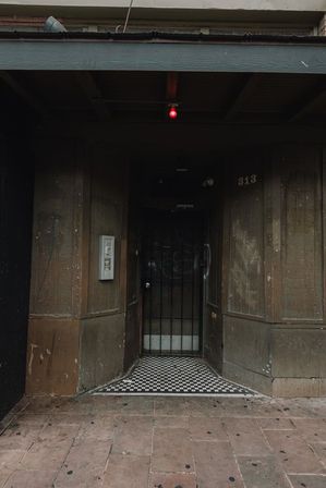 Urban storefront entrance with a black metal security gate, worn brown wood facade, black-and-white checkerboard tile threshold, small red hanging bulb above, and scuffed sidewalk.