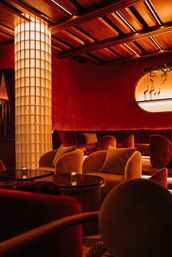 Intimate moody red cocktail lounge interior with plush velvet armchairs and curved banquette, glowing grid-textured column, warm wood-beamed ceiling, candlelit tables and backlit oval wall niche with flamingo silhouettes.