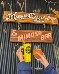 Rustic corrugated wall with wooden “Margarita Bar & Mimosa Bar” signs; two hands hold a bright yellow cocktail mug and a mimosa wine glass — brunch cocktails.