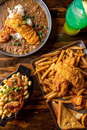 Overhead shot of a Southern seafood feast: crispy battered catfish and French fries with toasted bread, Cajun gumbo with rice and fried shrimp, creamy mac and cheese topped with grilled shrimp, and a green cocktail.